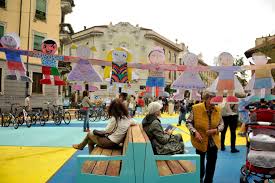 Piazza pedonale colorata, con panchine, persone sedute e giochi per bambini.